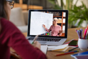 A person participates in an online learning session, watching a teacher explain concepts on a whiteboard via a laptop. The teacher is gesturing while speaking, and the student is taking notes with various stationery items spread out on the desk