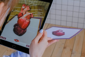 A person is holding a tablet that displays a 3D augmented reality model of a human heart. The model is superimposed over an image of a heart on a piece of paper
