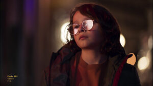 A girl sat in a dimly lit room with light bouncing off of her glasses