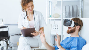 A patient sat in bed with a VR headset on. A smiling medical professional sits besides him on a chair with a notepad and a pen