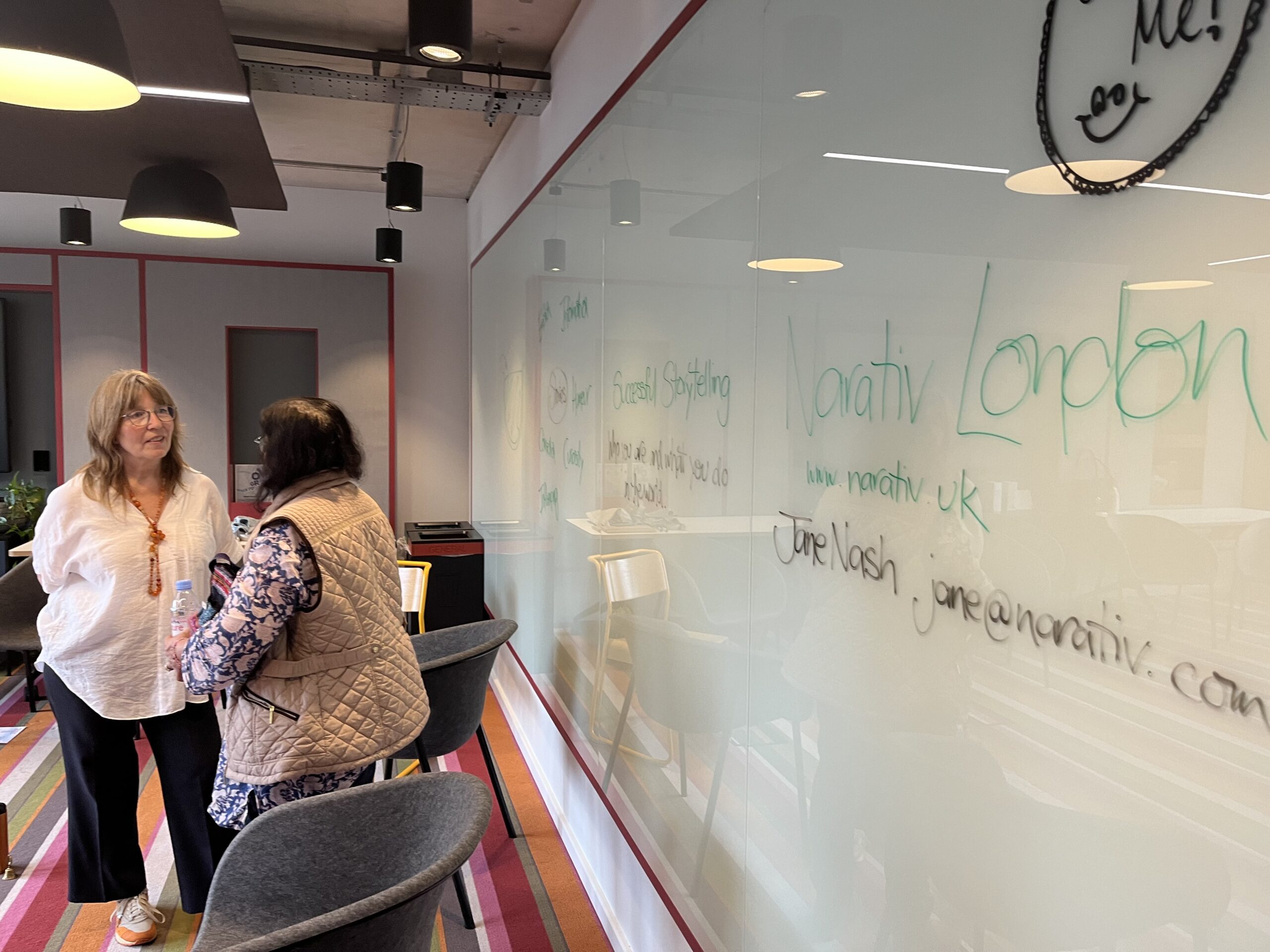 Two women talking in front of a glass whiteboard in a meeting room. The whiteboard has notes written in green and black marker, including 'Narativ London,' 'Successful Storytelling,' and contact information for Jane Nash.