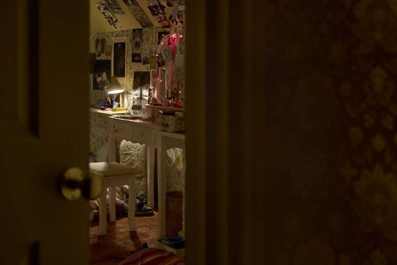 A dimly lit bedroom seen through a partially open door. The room features a vanity desk with a mirror and red string lights, a stool, and various personal items. The walls are covered with patterned wallpaper and decorated with posters.