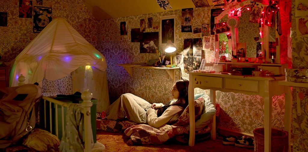 A dimly lit, cozy bedroom with a young woman lying on a mattress on the floor. The room is decorated with patterned wallpaper, posters, and various personal items. A tent made of white fabric with colourful lights is set up in one corner. A desk with a mirror and red string lights is against a wall.