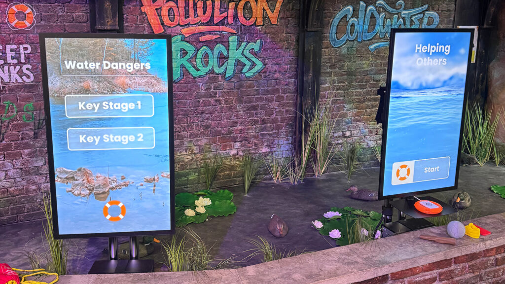 Interactive immersive technology exhibition setup with two touchscreen displays showing a water safety learning experience. One screen offers “Water Dangers” with Key Stage options, while the other displays “Helping Others” with a start button. The scene includes a themed environment with reeds, water features, and graffiti-style wall graphics.