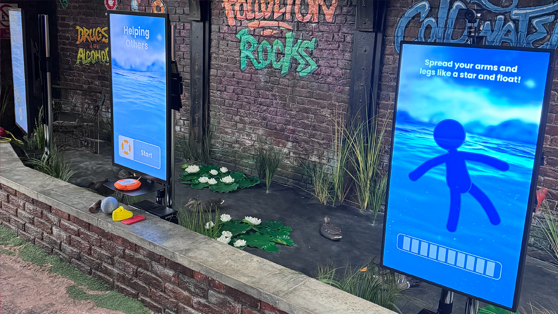 Interactive water safety installation with multiple vertical touchscreen displays in a themed environment featuring reeds, water plants, and graffiti-style walls. One screen shows a floating safety instruction—“Spread your arms and legs like a star and float!”—while another displays a “Helping Others” start screen.