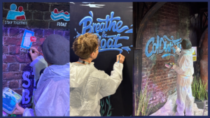 Artist hand-painting water safety graffiti including “Breathe & Float” and “Cold Water” messages on a themed brick wall during installation setup